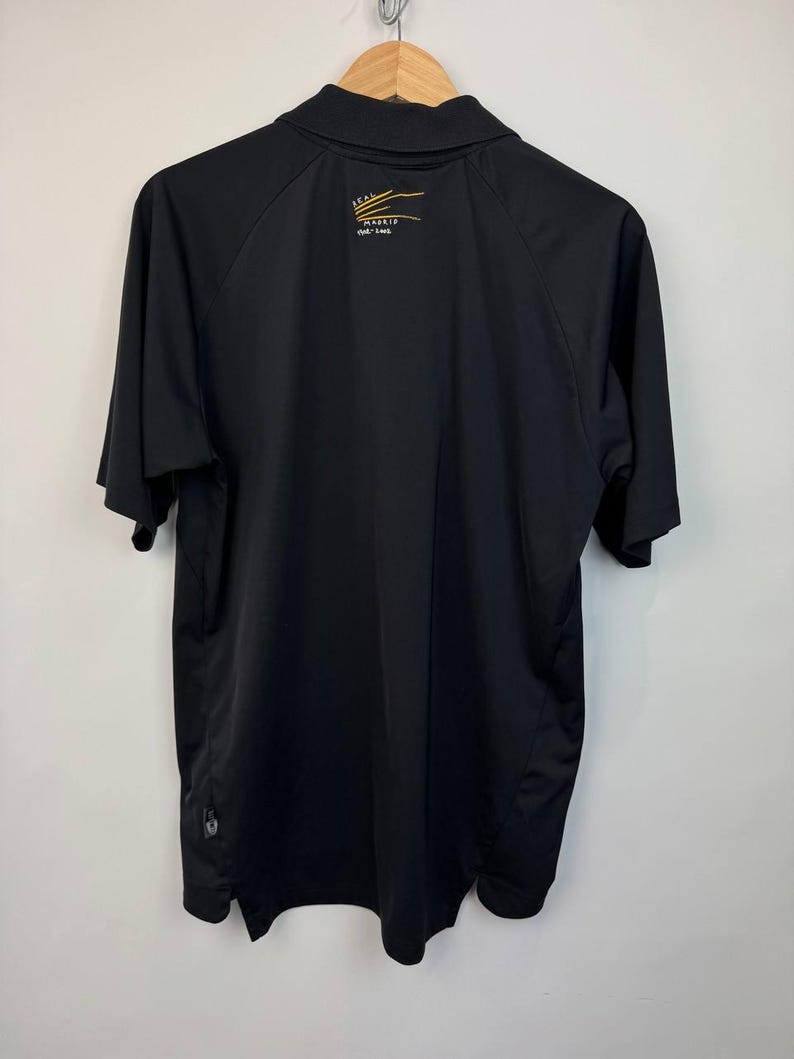 May include: Black polo shirt with short sleeves, hanging on a wooden hanger. The back of the shirt features a small gold and white logo with the text "REAL MADRID" and a smaller line of text below it. The shirt is made of a smooth, possibly athletic material.