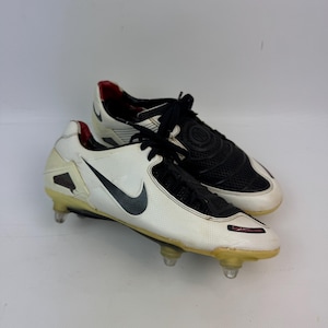 May include: Pair of white and black Nike soccer cleats with black laces and a black Nike swoosh. The cleats have clear studs and red accents on the heel and tongue. The soccer cleats are on a white background.