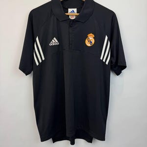 May include: Black Adidas polo shirt with white stripes on the sleeves and the Adidas logo on the chest. The shirt features a Real Madrid crest on the right chest. The shirt has a collared neckline and short sleeves.