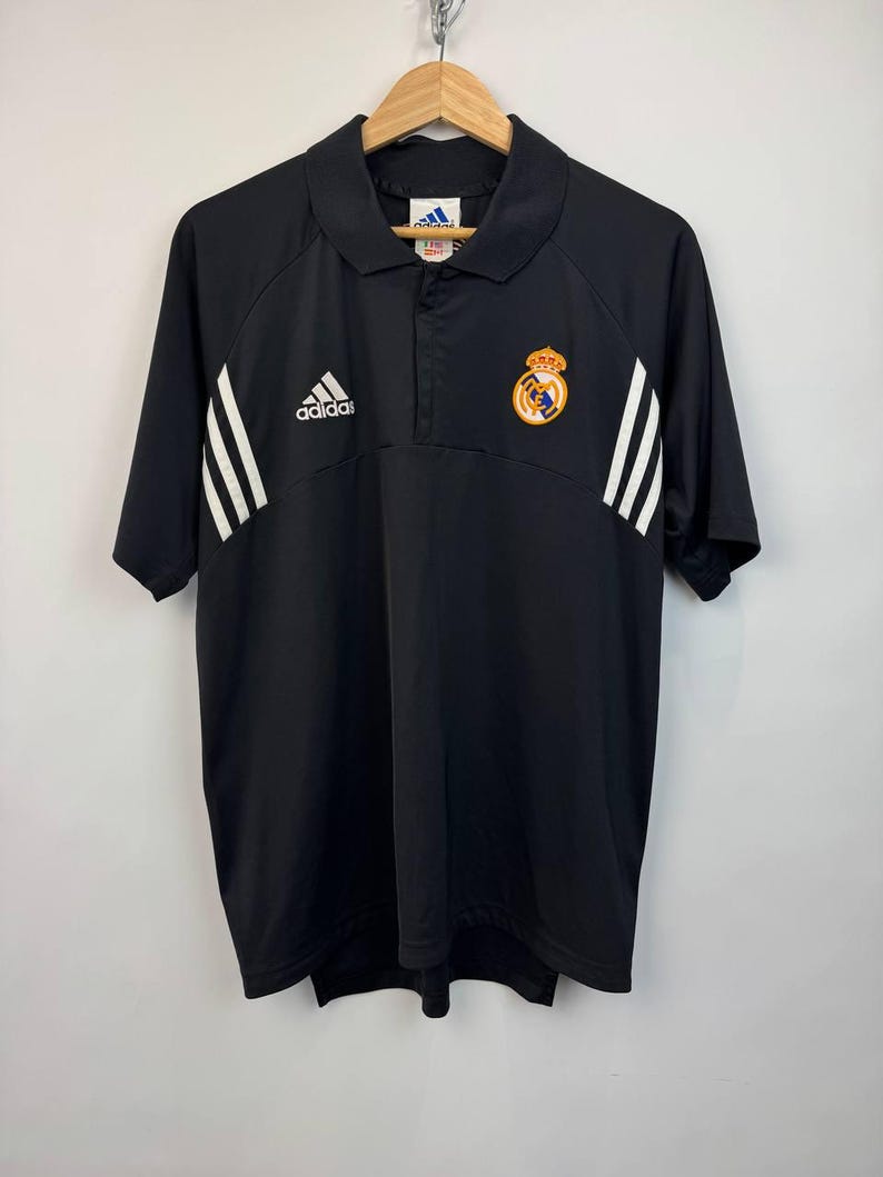 May include: Black Adidas polo shirt with white stripes on the sleeves and the Adidas logo on the chest. The shirt features a Real Madrid crest on the right chest. The shirt has a collared neckline and short sleeves.