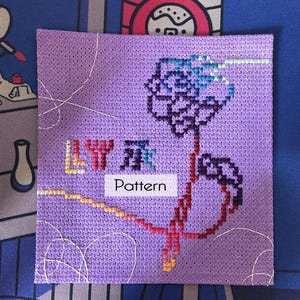 May include: A square, cross-stitched pattern on lavender fabric. The design features a stylized flower in shades of blue, purple, and red. The word "Pattern" is printed on a white rectangle. The text "LY" and Chinese characters are also visible.