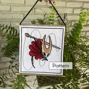 Pode incluir: Padrão de ponto cruz com um personagem com capa vermelha, cabeça bege e agulha. O desenho está em um fundo branco com costuras cinzas e a palavra "Pattern". A obra de arte é emoldurada com cavilhas de madeira e um cordão preto para pendurar.