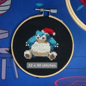 Pode incluir: Bordado em ponto cruz de uma personagem de desenho animado usando um gorro de Papai Noel, em um bastidor de madeira sobre um fundo preto. A personagem é azul, branca e marrom. O texto "32 x 30 pontos" está na parte inferior.
