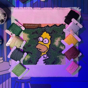 Puede incluir: Punto de cruz de Homer Simpson con carretes de hilo verde y amarillo. El punto de cruz presenta a Homer Simpson con una cara amarilla y una camisa blanca. Los carretes de hilo son de varios colores, incluyendo verde, amarillo, rosa y negro.