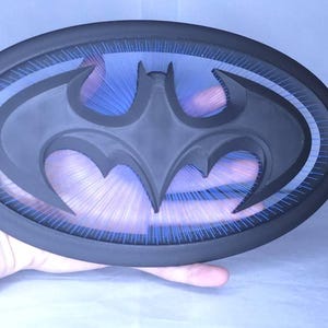 May include: A black oval-shaped wall plaque featuring a central cutout of the Batman bat symbol. The design includes a translucent blue backing with fine, radiating lines that create a sunburst effect behind the black bat silhouette.