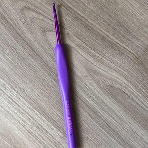 May include: A purple crochet hook, marked as 2.50 mm. It features a rounded handle and a thin, metallic hook. The tool is for yarn crafts, set against a wooden surface.
