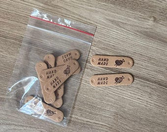 Może przedstawiać: Brązowe, owalne etykiety z napisem "HAND MADE" i motywem przędzy i igły do robienia na drutach. Niektóre etykiety są w przezroczystej plastikowej torebce. Idealne do rękodzieła.
