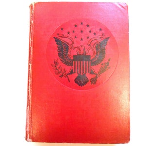 May include: A red book with a black emblem featuring an eagle, stars, and stripes. The book's cover is a solid red colour, and the emblem is in a circular shape. The book appears to be vintage.