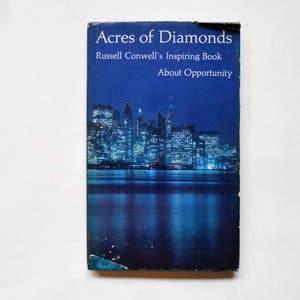 May include: A vintage book titled "Acres of Diamonds" by Russell Conwell. The cover features a nighttime cityscape with illuminated buildings reflected in the water, set against a dark blue background. The book is slightly worn.