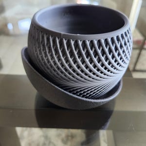 May include: A grey, 3D-printed plant pot with a matching saucer. The pot has a spherical shape with a lattice design, and the saucer is a simple, shallow dish. The items are made of a matte material.