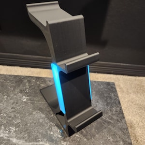 May include: A black, modern-style phone stand with a built-in blue LED light. The stand is made of a matte material and has a curved design to hold a phone or tablet. It sits on a marble-patterned surface.