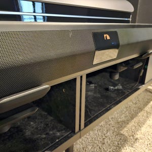 Puede incluir: Una barra de sonido gris oscuro con un panel frontal perforado y una pequeña pantalla. La barra de sonido está montada en un mueble gris oscuro con puertas de vidrio y patas con punta dorada. El mueble se encuentra sobre una alfombra clara.