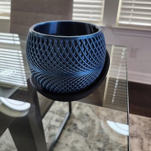 Pode incluir: Um vaso azul, impresso em 3D, com um design vazado único. O vaso repousa sobre um porta-copos redondo e escuro. O design apresenta um padrão repetitivo de linhas curvas, criando uma textura visualmente interessante. O vaso está vazio.