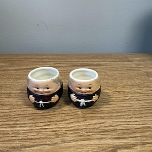 Puede incluir: Dos hueveras de cerámica con forma de monjes de dibujos animados. Las hueveras tienen un interior blanco, túnicas marrones y caras sonrientes. Las hueveras están sobre una superficie de madera.