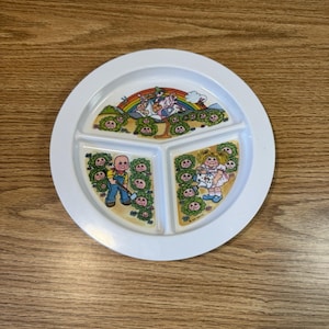 Könnte beinhalten: Weißer Teller mit Unterteilung und farbenfrohem Cabbage Patch Kids-Design. Der Teller hat drei Abschnitte und zeigt Illustrationen der Cabbage Patch Kids mit einem Regenbogen und anderen Figuren. Der Teller befindet sich auf einer Holzoberfläche.