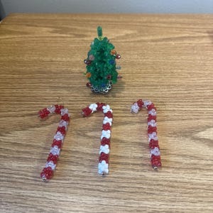 Puede incluir: Un árbol de Navidad verde con cuentas con adornos coloridos y tres adornos de bastón de caramelo con cuentas. Los bastones de caramelo son rojos y blancos, con detalles plateados. Los adornos están sobre una superficie de madera.