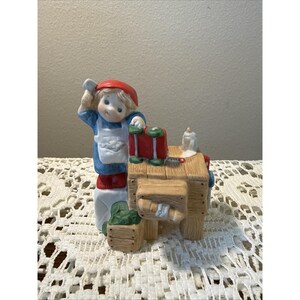 May include: A ceramic figurine of a child wearing a red hat, blue shirt, and white apron, standing beside a wooden workbench. The workbench features a red toolbox, a small white pitcher, and a green crate, all rendered in a painted finish.