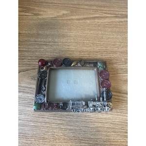 Op de afbeelding: Een rechthoekige fotolijst met een bruine rand en een treinthema. De lijst is versierd met diverse versieringen, waaronder sleutels, tandwielen en rode en groene kralen. De lijst is ongeveer 15 cm breed en 10 cm hoog.