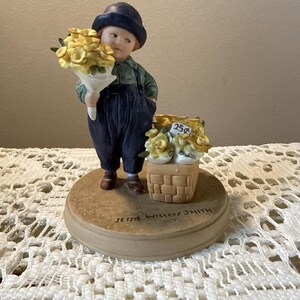 May include: A ceramic figurine of a child wearing a dark blue hat, green shirt, and dark blue overalls. The child holds a bouquet of yellow flowers and stands next to a woven basket filled with yellow flowers marked with a 25 cent price tag.
