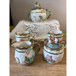 Puede incluir: Un juego de té de cerámica que incluye una tetera, una lechera, un azucarero con tapa de conejo y dos tazas con platillos. Cada pieza es blanca con motivos florales en relieve, detalles de enredaderas verdes y bordes en tono dorado.