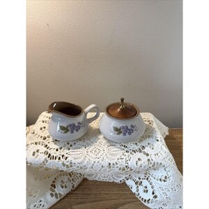 Peut inclure: Un pot à lait et un sucrier avec couvercle en céramique, présentant une base blanche ornée d'un motif de raisins violets et de feuilles vertes. Le pot à lait a un bord intérieur marron, et le sucrier possède un couvercle en bois avec un bouton en métal.