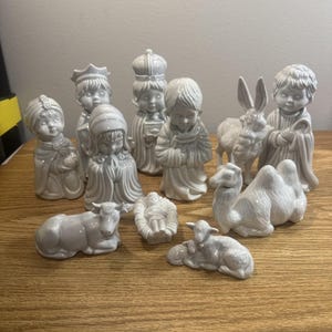 May include: A collection of ceramic nativity figurines in a soft gray color. The set includes figures of the three wise men, Mary, Joseph, baby Jesus, animals, and an angel. The figurines are arranged on a wooden surface.