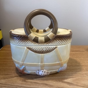 May include: A ceramic basket-shaped container with a brown handle and lid. The container is cream-colored with brown accents, including a woven pattern around the rim and straps. Seashell and starfish details adorn the front.