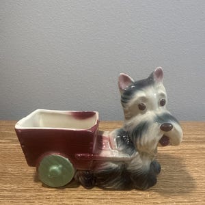 Può includere: Una fioriera in ceramica a forma di cane Scottish Terrier che tira un carretto rosso. Il cane ha pelo nero, bianco e grigio con orecchie rosa. Il carretto ha un interno bianco e ruote verdi. Un oggetto decorativo.
