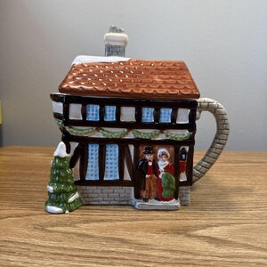 Puede incluir: Una tetera de cerámica con forma de casa de dos pisos con tejado marrón y chimenea. La casa tiene fachada de piedra, vigas marrones y ventanas azules. Un pequeño árbol de Navidad verde y una pareja con ropa de época están al frente.