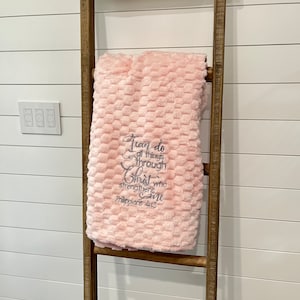 May include: A soft, pink blanket with a textured, honeycomb pattern is draped over a wooden ladder. The blanket features embroidered text that reads "I can do all things through Christ who strengthens me, Philippians 4:13."