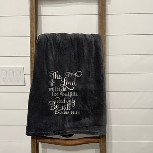 May include: A dark gray fleece throw blanket with white embroidered text that reads "The Lord will fight for you; you need only be still."  Exodus 14:14