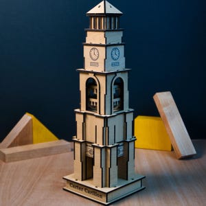 May include: A wooden model of a clock tower with intricate laser-cut details. The tower features a square base, multiple levels, arched openings, and a peaked roof. The clock faces show the time. The base reads "Carter Carillon".