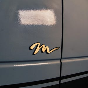 以下が含まれることがあります： スタイリッシュなデザインの金色の文字「M」のエンブレム。明るいグレーの車両パネルに取り付けられています。パネルには黒いレーシングストライプと垂直の継ぎ目があります。エンブレムには影の効果があります。