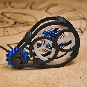 May include: A 3D-printed gear mechanism in black, white, and blue. The design incorporates interlocking gears and circular elements. The gears are blue, with white and black components. The object is placed on a tan background, with a blueprint design.