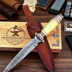 May include: A handcrafted dagger with a patterned blade and a light yellow handle, resting in a brown leather sheath. The dagger is displayed on a wooden box with the text "TRUSTEDKNIVESBYAWAIS Etsy Shop". The overall aesthetic is rustic and artisanal.
