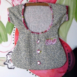 May include: A gray sleeveless baby onesie with a pink and white floral print around the neckline. The onesie has a small pocket on the front with a pink and white floral print. There are three white buttons down the front of the onesie.