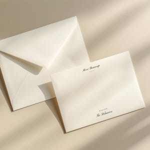 Op de afbeelding: Ivoor briefpapier set met een gevouwen envelop en een kaart. De kaart heeft de woorden "Merci Beaucoup" en "With Love The Delauriers" in zwarte letters. De set staat op een neutrale achtergrond met zachte schaduwen.