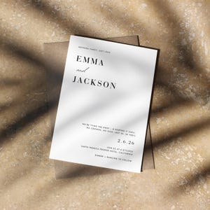 May include: A white wedding invitation with the names "EMMA and JACKSON" in elegant black font. The invitation includes the date "2.6.26" and event details. The invitation is on a brown envelope, with shadows.