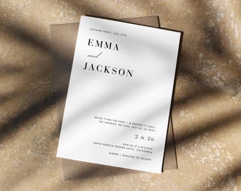 May include: A white wedding invitation with the names "EMMA and JACKSON" in elegant black font. The invitation includes the date "2.6.26" and event details. The invitation is on a brown envelope, with shadows.
