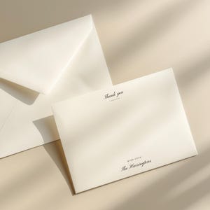 Peut inclure: Ensemble de papeterie ivoire comprenant une carte pliée et une enveloppe. La carte porte l'inscription "Thank you" en écriture élégante, avec "With Love" et un nom de famille en dessous. L'ensemble est posé sur une surface neutre.