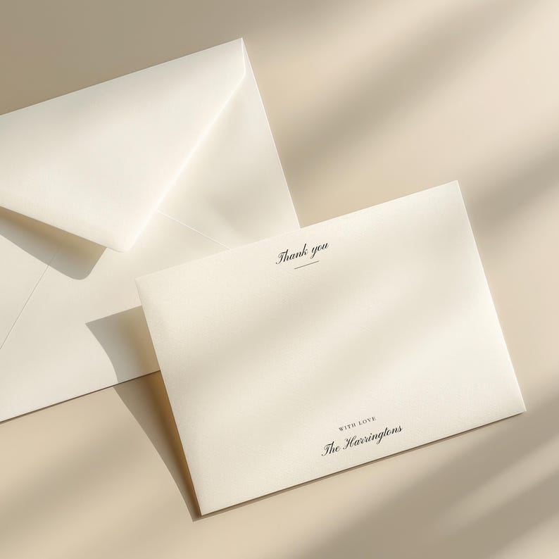 May include: Ivory stationery set featuring a folded card and envelope. The card reads "Thank you" in elegant script, with "With Love" and a family name below. The set is arranged on a neutral surface, with soft shadows.