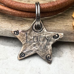 May include: A star-shaped metal pet tag with a hammered texture and dark spiral accents at each point. The name Lucy is engraved in the centre of the tag. The tag is attached to a silver-toned metal split ring, which is threaded onto a brown leather cord.