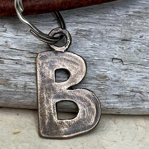 May include: A bronze-colored metal keychain charm in the shape of the letter "B". The charm is attached to a silver-colored keyring. The background is a weathered, gray wooden surface.