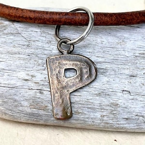 May include: A bronze-toned metal pendant shaped like the capital letter P with a hammered, textured surface. The pendant is attached to a silver-colored metal jump ring, which is threaded onto a thick, round, brown leather cord.