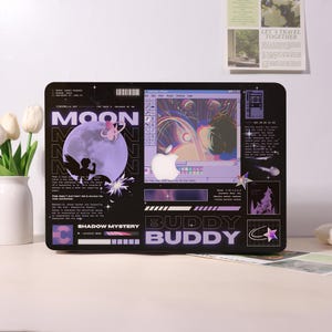 May include: Black laptop skin with a graphic design featuring a purple moon, an angel, and the text "MOON". The design also includes a computer interface with an Apple logo and the words "BUDDY BUDDY".