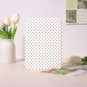 May include: A white rectangular object with a black polka dot pattern. The object is standing upright on a light-colored surface. A white vase with tulips is to the left.