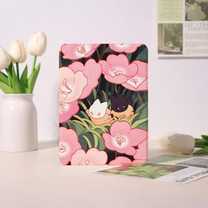 May include: A tablet case featuring a floral design with pink flowers and green leaves. Two cartoon cats, one white and one black, are nestled among the flowers. The case has a glossy finish and a rectangular shape.