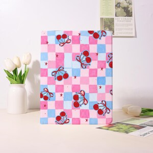 May include: A rectangular, patterned item featuring a checkerboard design in pink and blue squares. Adorning the surface are illustrations of red cherries tied with pink bows and small red stars. The item is likely a decorative piece or accessory.