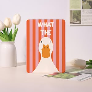 Può includere: Una custodia rettangolare con un design a forma di anatra dei cartoni animati. Lo sfondo presenta strisce verticali arancioni e rosa. Le parole "WHAT THE" sono stampate in bianco sopra l'illustrazione dell'anatra.