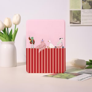 May include: A pink tablet case with a red and pink striped bottom half. The top half features illustrations of a frog, a zebra, a duck, and a dog on a scooter. The case is standing on a white surface.